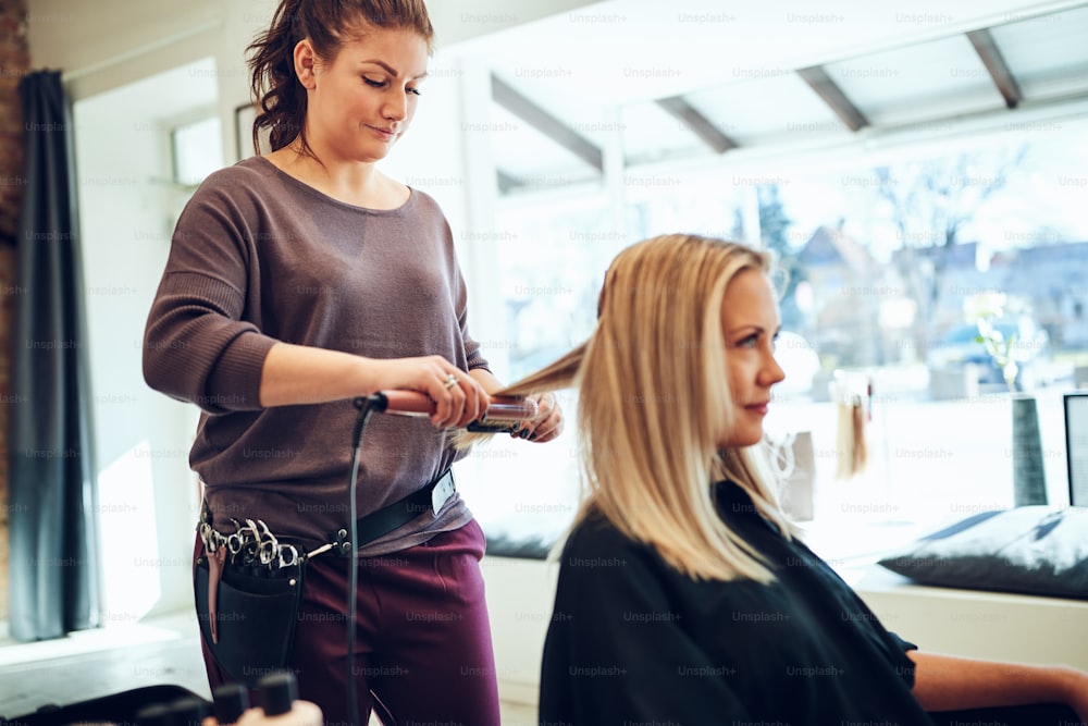 Smiling young blonde woman sitting in a chair in a salon having her hair straightened by her hairstylist