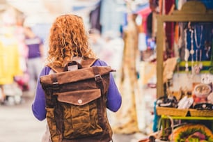 Blonde curly hair woman backpacker traveler viewed from rear at used market enjoying the shopping and alternative vacation - leather backpack view for travel concept