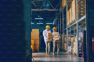 Two blue collar workers in white uniforms and with yellow helmets on heads relocating heavy boxes in warehouse.