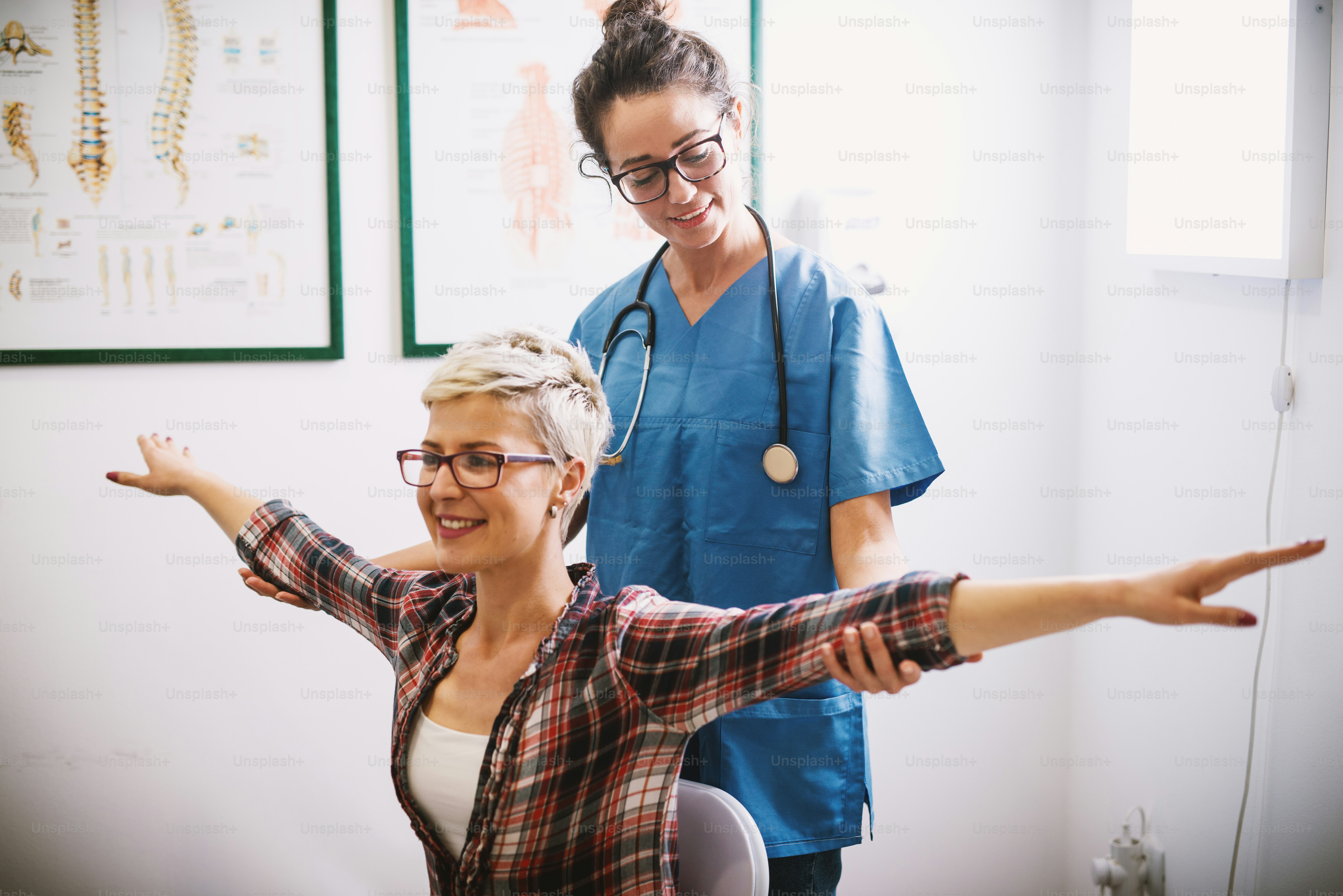 Infermiera professionale di mezza età focalizzata che tiene e controlla i gomiti della paziente donna mentre è seduta su una sedia di fronte a braccia aperte.