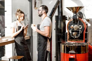 Couple of baristas talking during the coffee break standing together in the coffee shop with roaster machine on the background