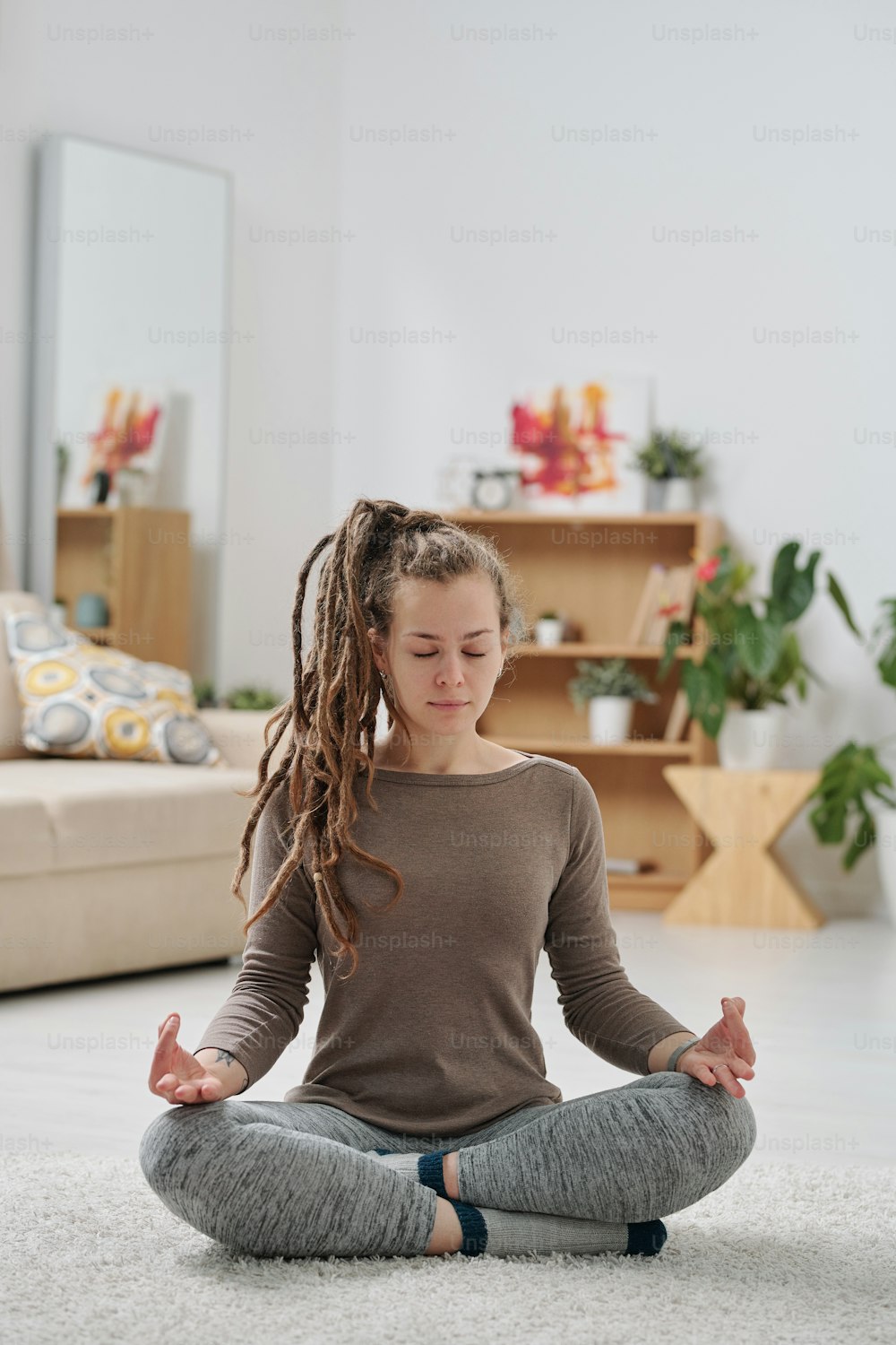 Hübsche junge aktive Frau mit Dreadlocks, die mit gekreuzten Beinen auf dem Boden sitzt und zu Hause Yoga praktiziert