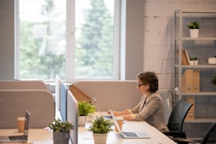 Serious busy young lady with short hair sitting at table in modern office and using laptop while analyzing sales forecast