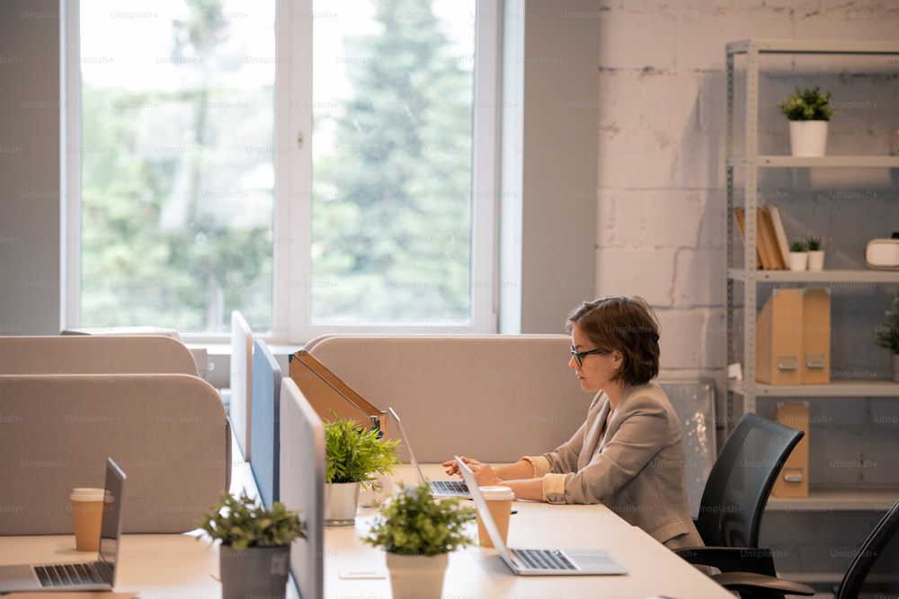 Serious busy young lady with short hair sitting at table in modern office and using laptop while analyzing sales forecast