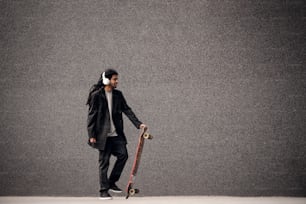 Stylish young dreadlocks hipster skater with headphones holding skateboard near the grey wall in a suit.