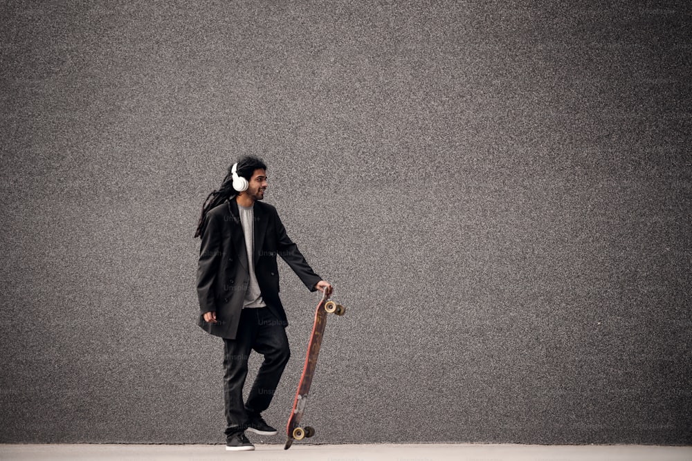 Giovane skater hipster elegante con i dreadlocks con le cuffie che tiene lo skateboard vicino al muro grigio in un vestito.
