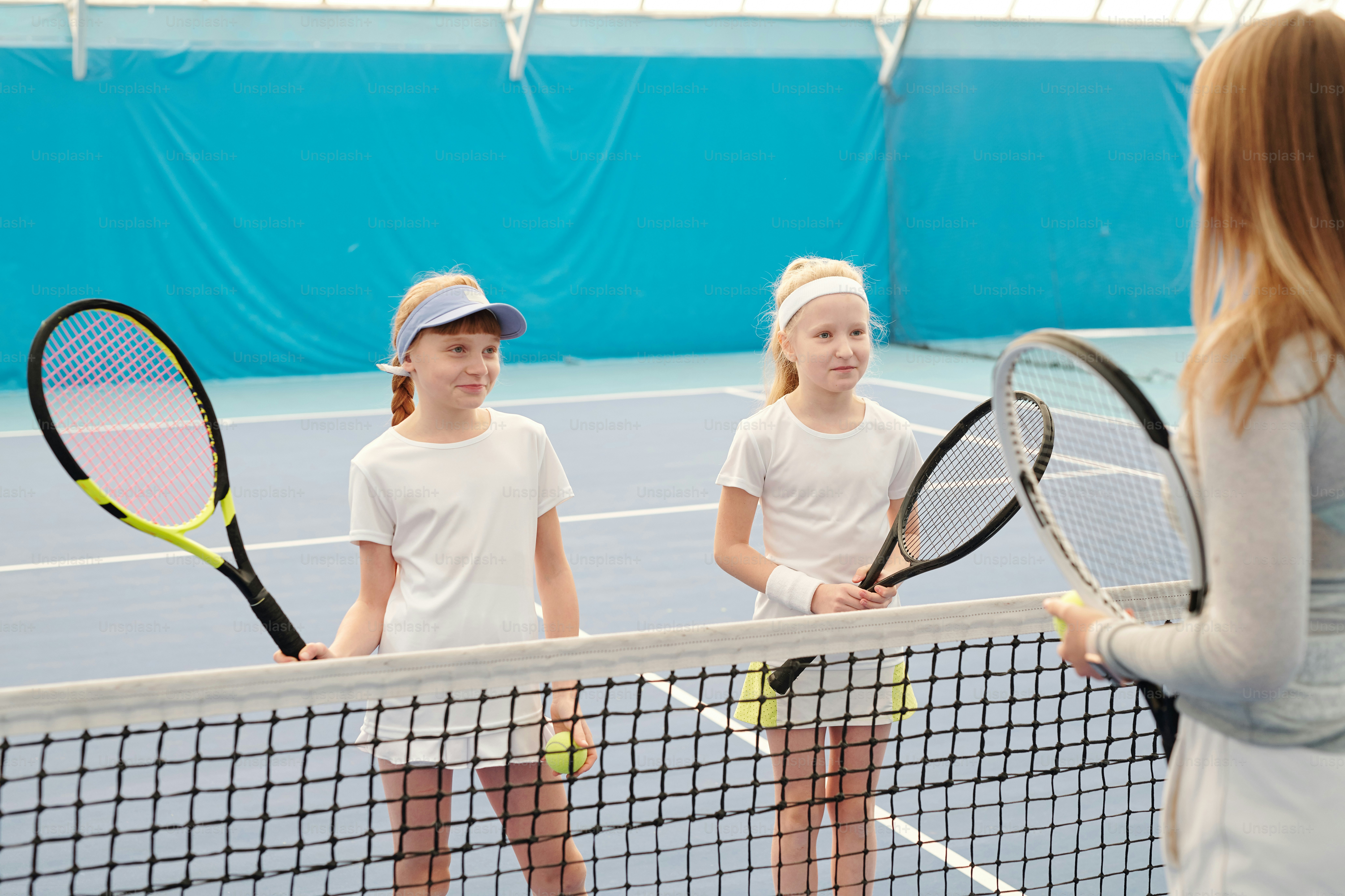 Two happy girls in white sportswear holding tennis rackets while standing by net in front of their instructor and listening to her explanations