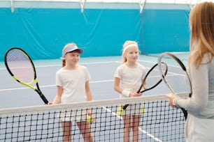 Two happy girls in white sportswear holding tennis rackets while standing by net in front of their instructor and listening to her explanations