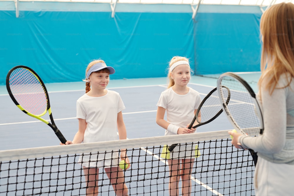 Due ragazze felici in abbigliamento sportivo bianco che tengono in mano racchette da tennis mentre sono in piedi vicino alla rete di fronte al loro istruttore e ascoltano le sue spiegazioni