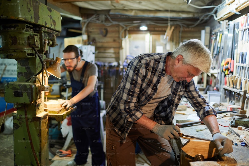 Maestro senior di dettaglio di lavorazione del legno con scalpello mentre il suo giovane apprendista lavora a macchina