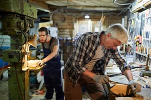 Maestro superior de carpintería procesando detalles con cincel mientras su joven aprendiz trabaja a máquina