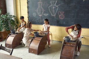 Gruppo di bambini in grembiuli che scolpiscono tazze di argilla su ruote di ceramica sotto il controllo dell'insegnante in classe