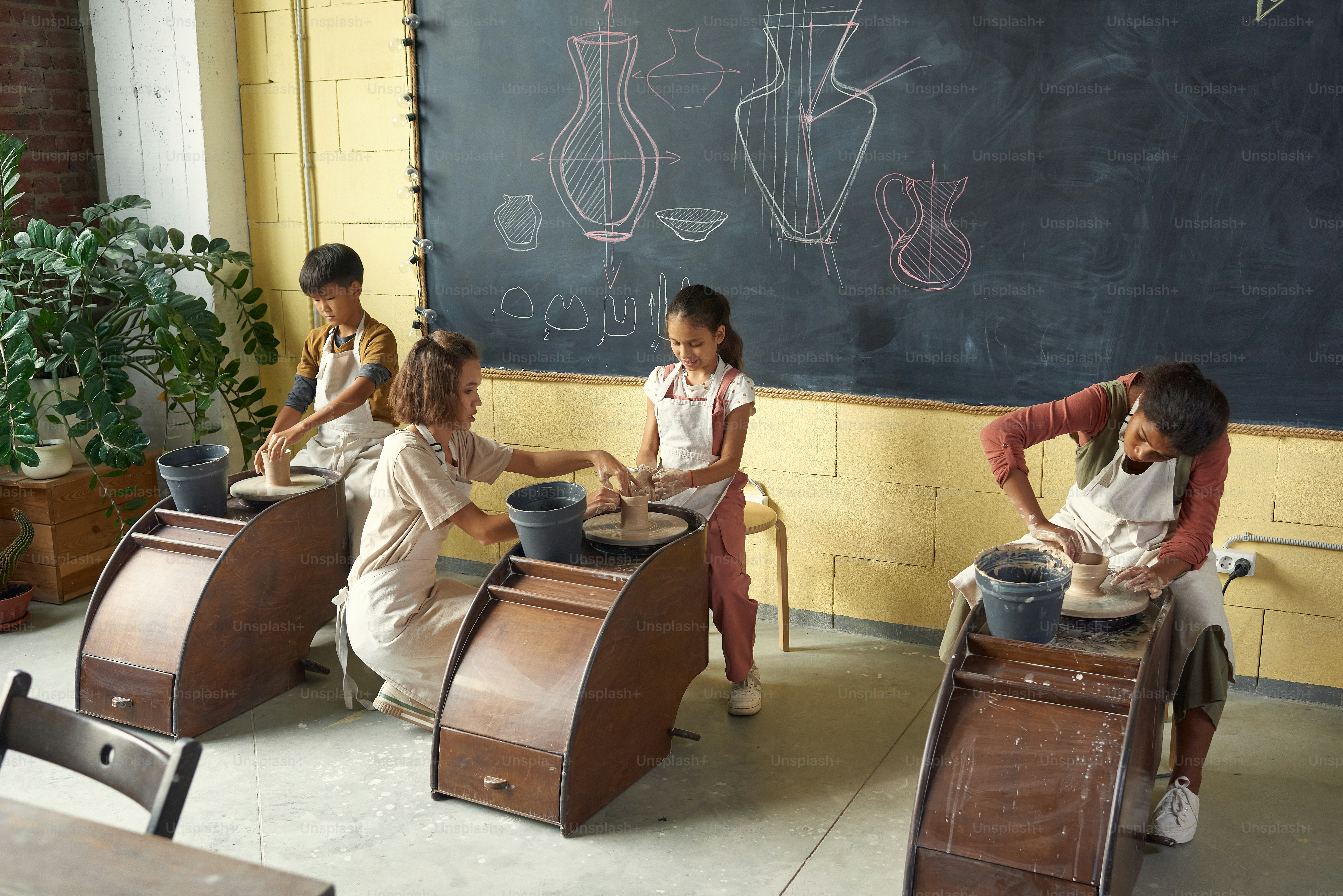 Group of kids in aprons sculpting clay cups on pottery wheels under control of teacher in classroom