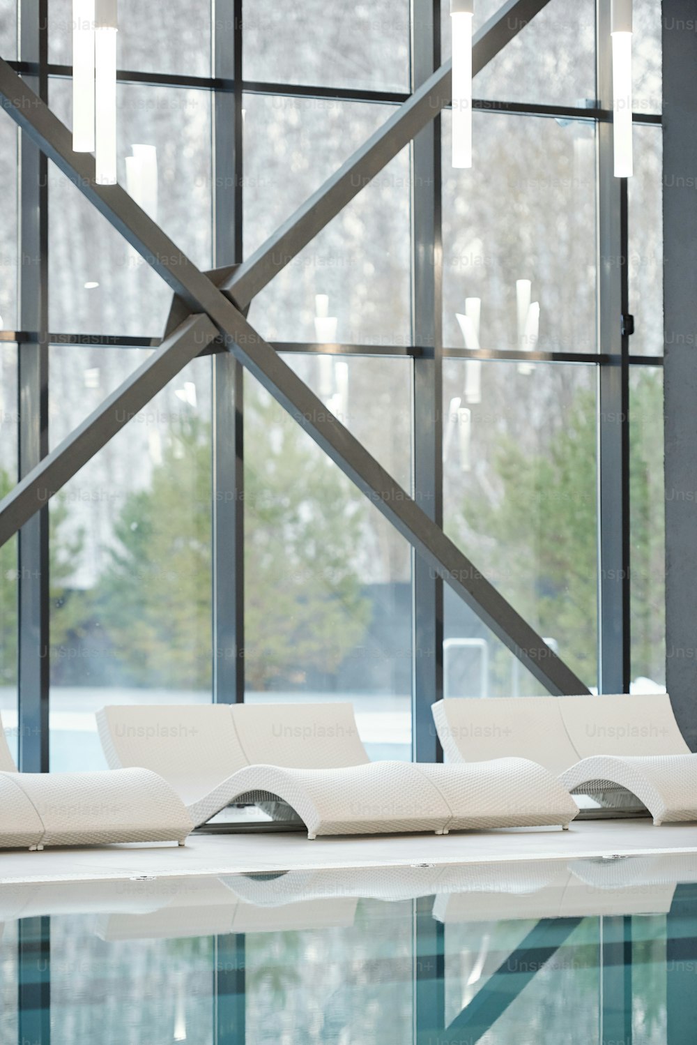 Fila de tumbonas blancas de pie a lo largo de grandes ventanales y piscina con agua transparente dentro de un moderno hotel, spa o centro de ocio