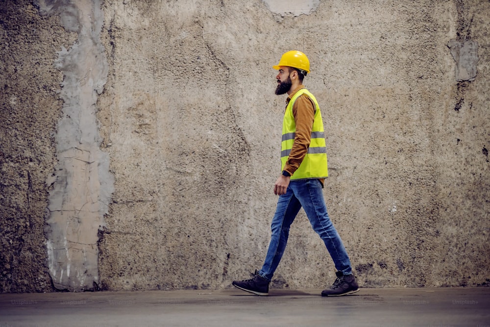 Side view of handsome hardworking bearded worker in vest, with safety helmet on head passing by concrete wall in building in construction process.