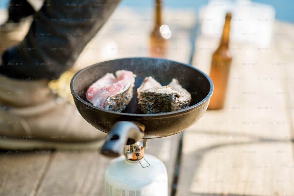 Freír dos filetes de pescado en el quemador durante el picnic con cerveza y aparejos de pesca al aire libre