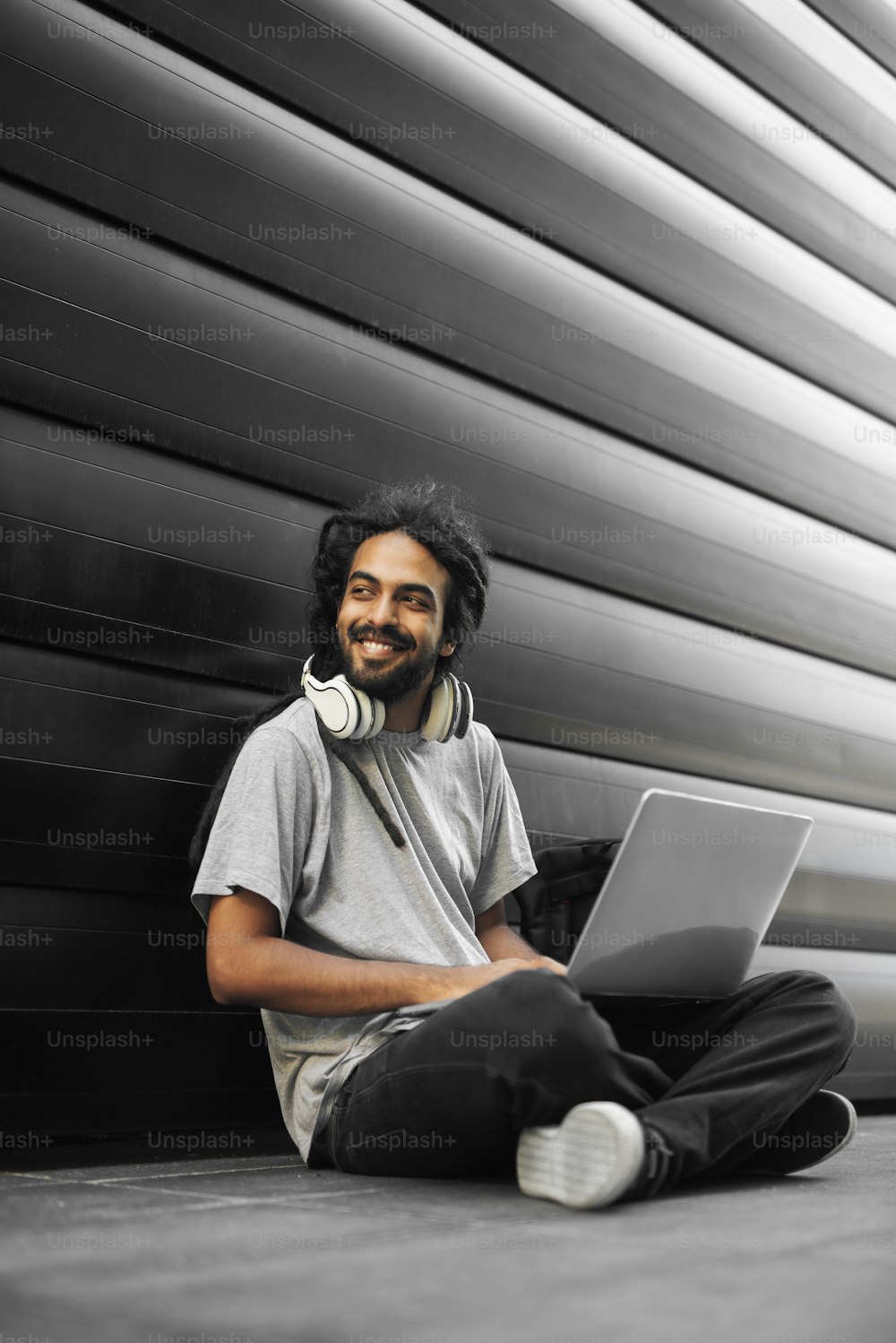 Lässiger indischer Hipster, der auf der Straße sitzt, auf dem Laptop tippt und zur Seite schaut.