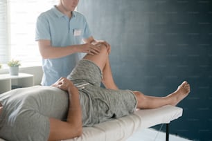 Male patient lying on medical couch while young physiotherapist massaging his bent left knee during rehabilitation course