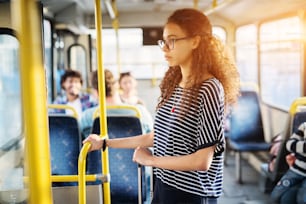 Young cute girl with curly hair is holding onto bar and her luggage while standing in a bus.