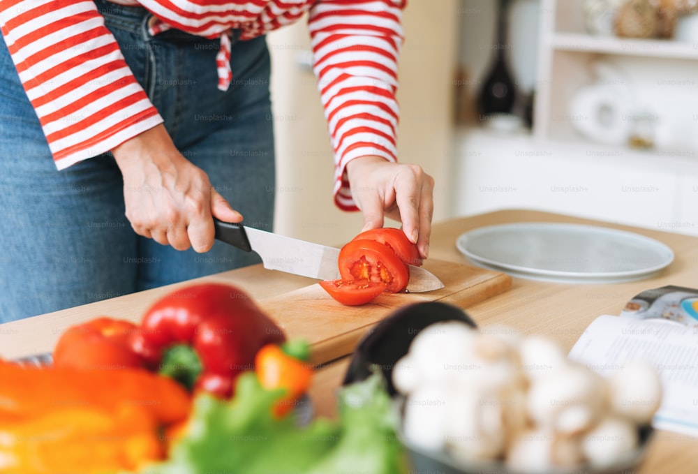 Fresh vegetables green salad, peppers, mushrooms and eggplant on kitchen table. Young woman plus size body positive in red longsleeve cooking using book with recipes on kitchen