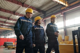 Confident factory workers in yellow hardhats walking together in industrial workshop and discussing work processes