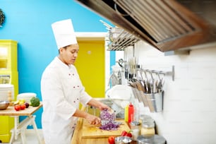 Joven chef cortando repollo morado fresco mientras está de pie junto a la mesa para preparar comida en la cocina