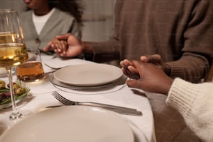 Young couple and their daughter holding by hands while sitting by served festive table and praying before dinner