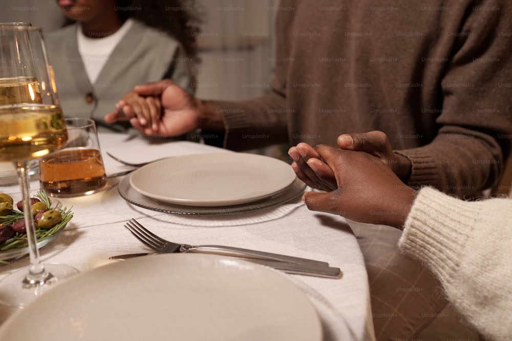 Pareja joven y su hija tomados de la mano mientras están sentados junto a la mesa festiva servida y rezan antes de la cena