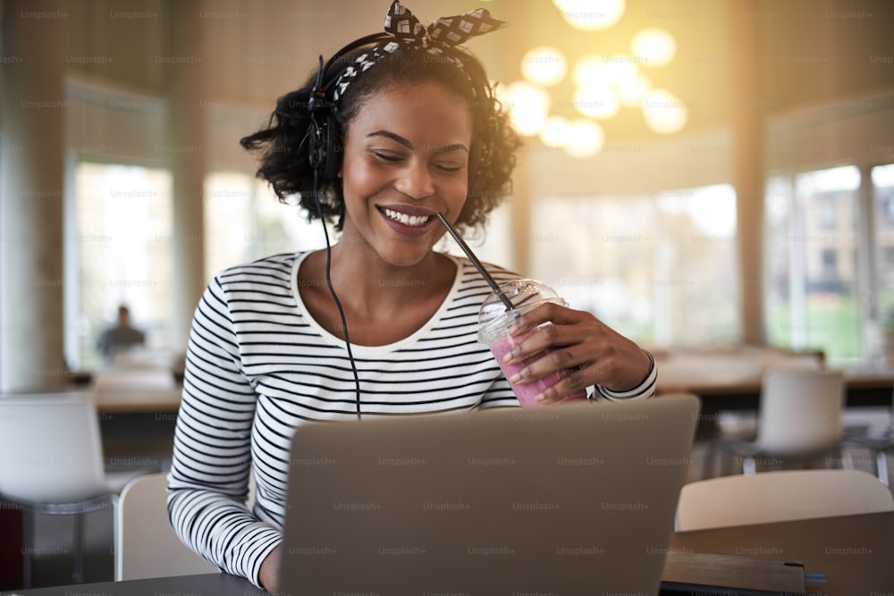 Joven estudiante universitario africano sonriente sentado en el campus trabajando en una computadora portátil y bebiendo un batido mientras escucha música con auriculares