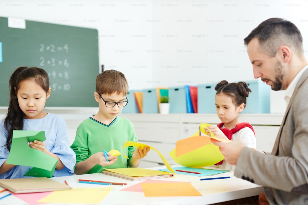 Niños de la escuela primaria sentados en el aula y haciendo origami con el maestro masculino mientras tienen una clase de arte después de la escuela