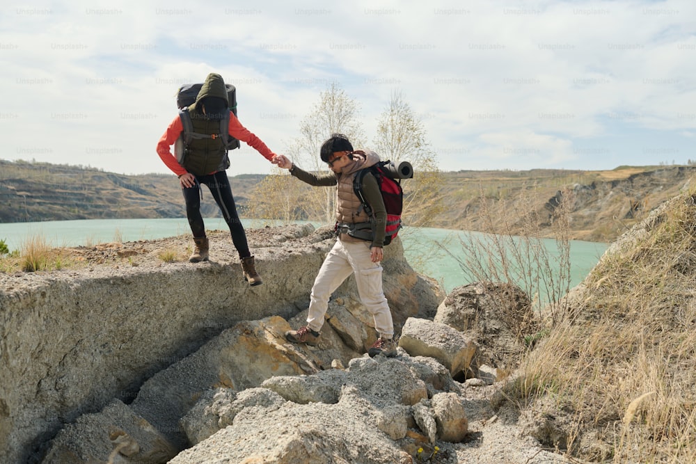 Horizontale Weitaufnahme von zwei jungen Frauen, die ein Wochenende zusammen beim Wandern um den Steinbruchsee verbringen, Frau hilft ihrer Freundin, den Felsen hinunter zu gehen