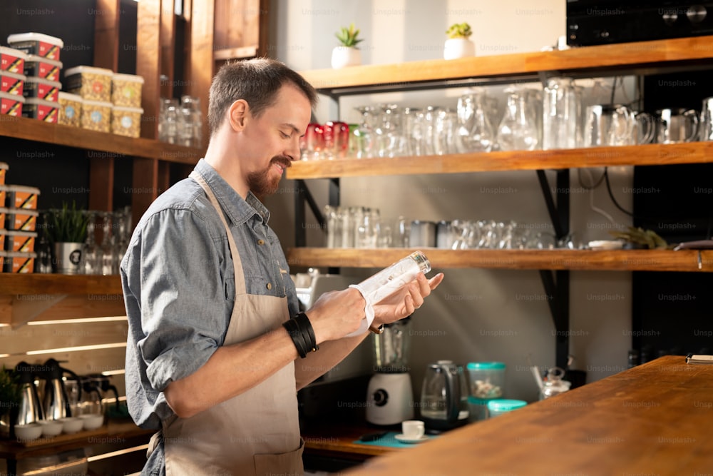 Happy young bearded waiter in apron wiping glass with napkin or towel while standing by counter among shelves with kitchenware
