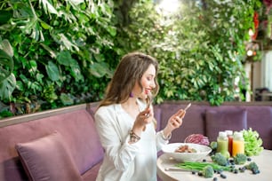 Mulher de negócios jovem comendo comida saudável sentada no restaurante vegetariano com a parede viva de plantas verdes no fundo