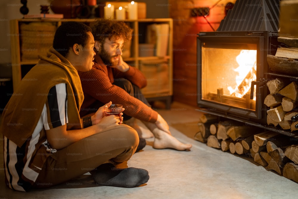 Two men sitting together by the burning fireplace at cozy home. Concept of homosexual relations and coziness on winter time. Idea of multinational gay families