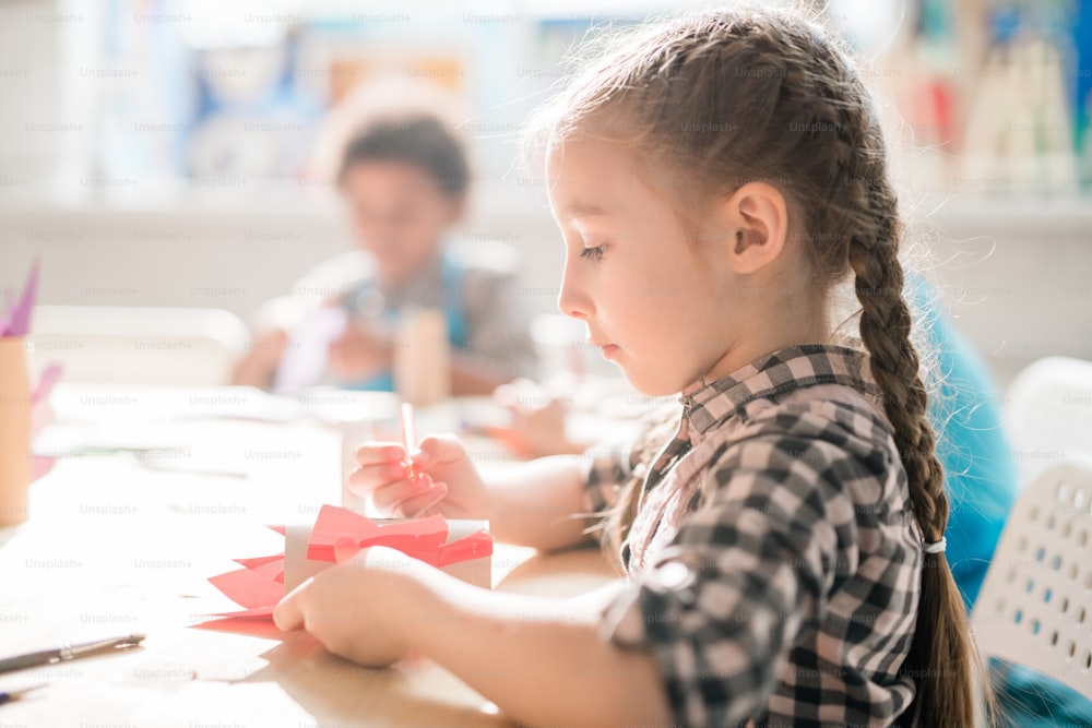 Creative schoolgirl sitting by desk and making decoration or toy at lesson during individual work