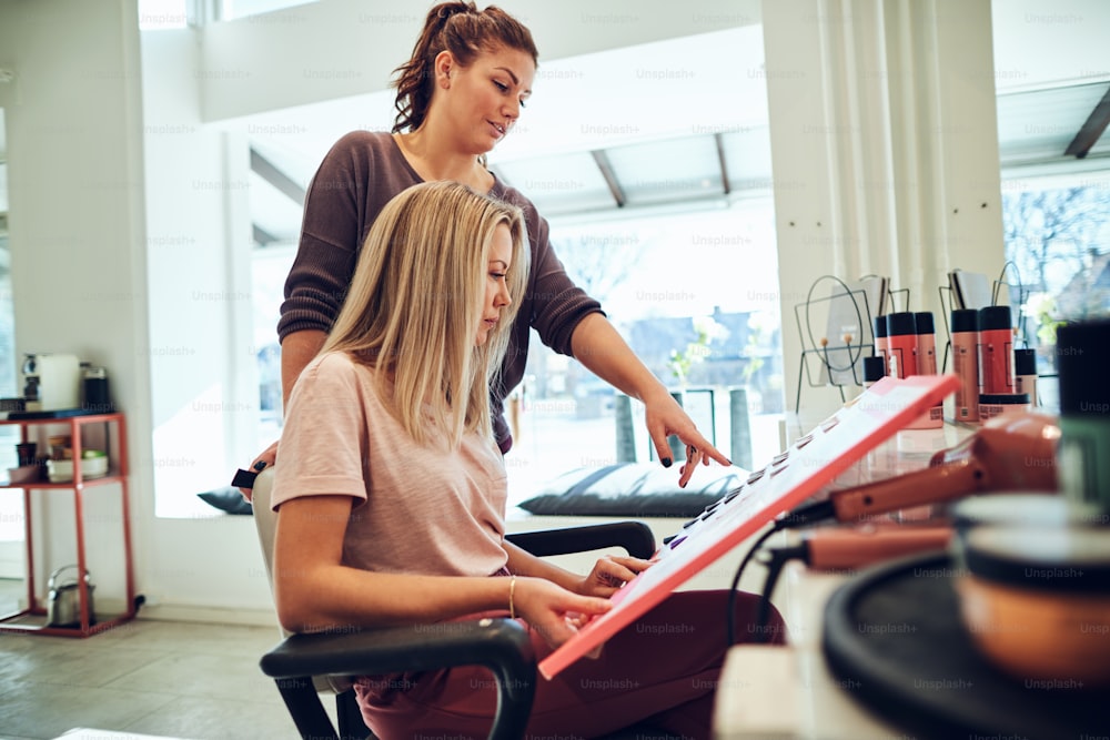 Young blonde woman looking through hair dye samples in a book with her hairstylist while sitting in a salon chair