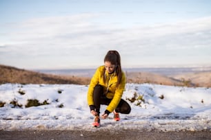화창한 아침에 이어폰으로 눈 덮인 겨울 도로에서 겨울 운동복에 신발 끈을 묶는 젊은 날씬한 운동 소녀.