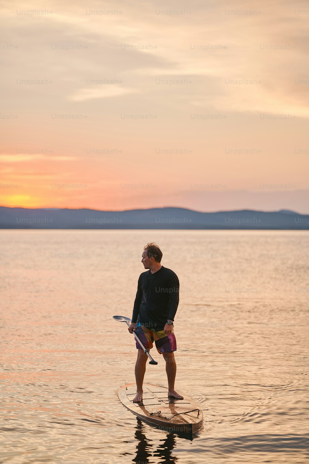 Full body shot of a man with paddle standing on wooden board in quiet water, surfing on sunset with scenic mountain view, looking away