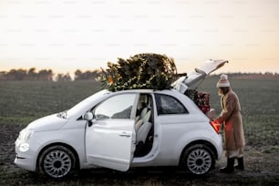Frau packt Geschenke ins Auto mit Weihnachtsbaum auf einem Dach in der Natur in der Abenddämmerung. Vorbereitung auf einen Neujahrsurlaub. Idee einer Weihnachtsstimmung. Frau mit Pelzmantel und Hut