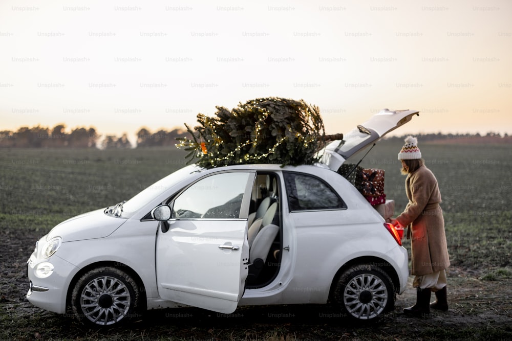 Frau packt Geschenke ins Auto mit Weihnachtsbaum auf einem Dach in der Natur in der Abenddämmerung. Vorbereitung auf einen Neujahrsurlaub. Idee einer Weihnachtsstimmung. Frau mit Pelzmantel und Hut