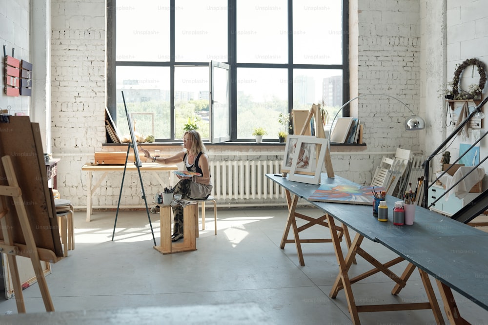 캔버스 앞에 앉아 넓은 스튜디오에서 그림을 그리는 여성 예술가