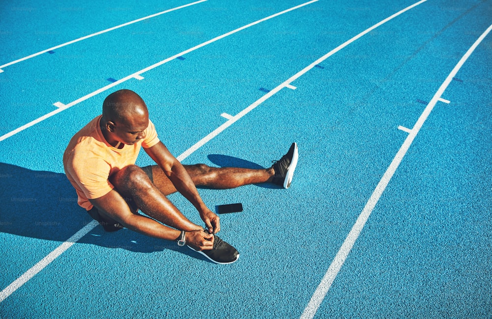 Giovane atleta maschio africano concentrato seduto da solo su una pista da corsa che si allaccia le scarpe prima dell'allenamento