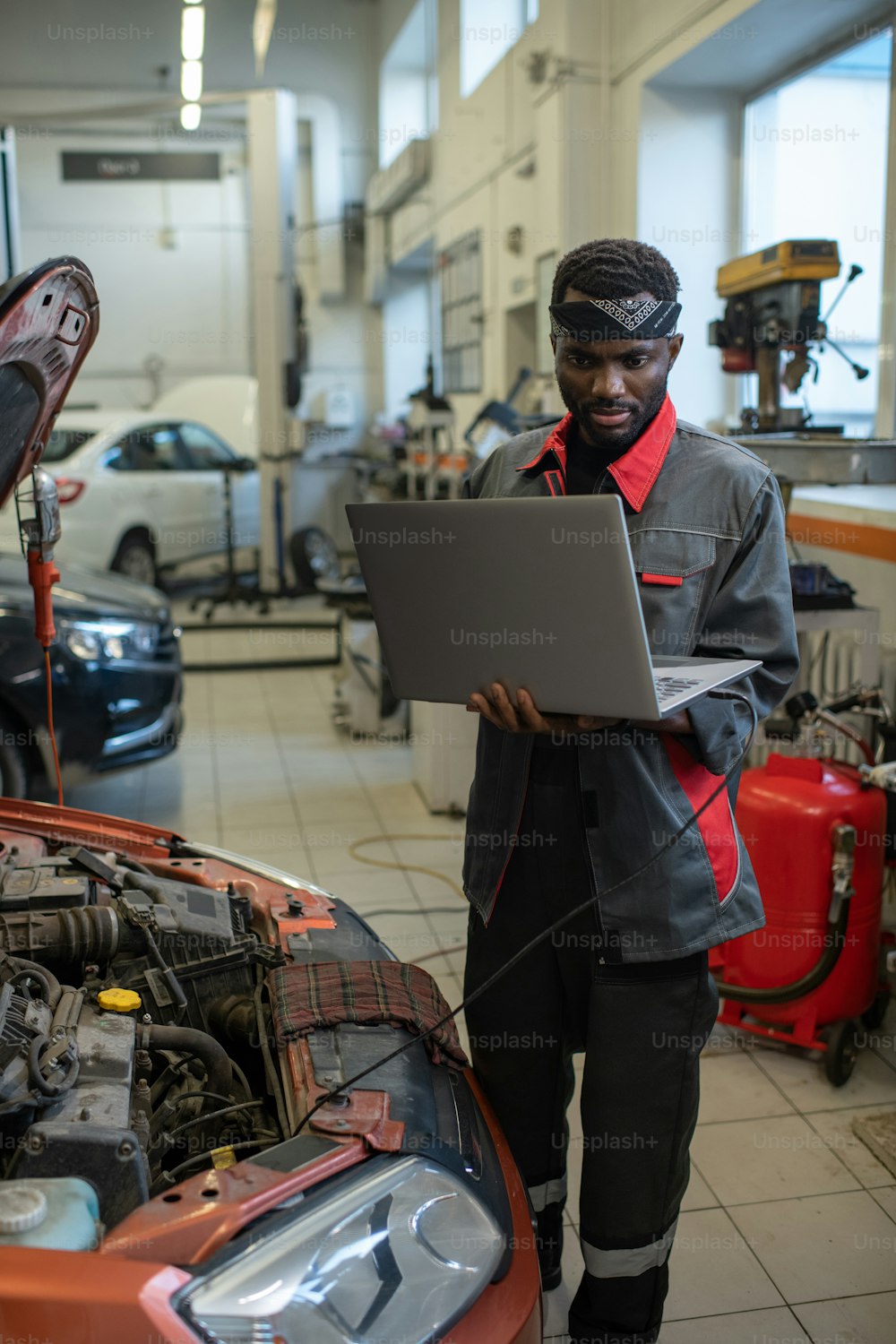 Vertical portrait of African-American mechanic standing by car in auto repair shop and using laptop while inspecting vehicle