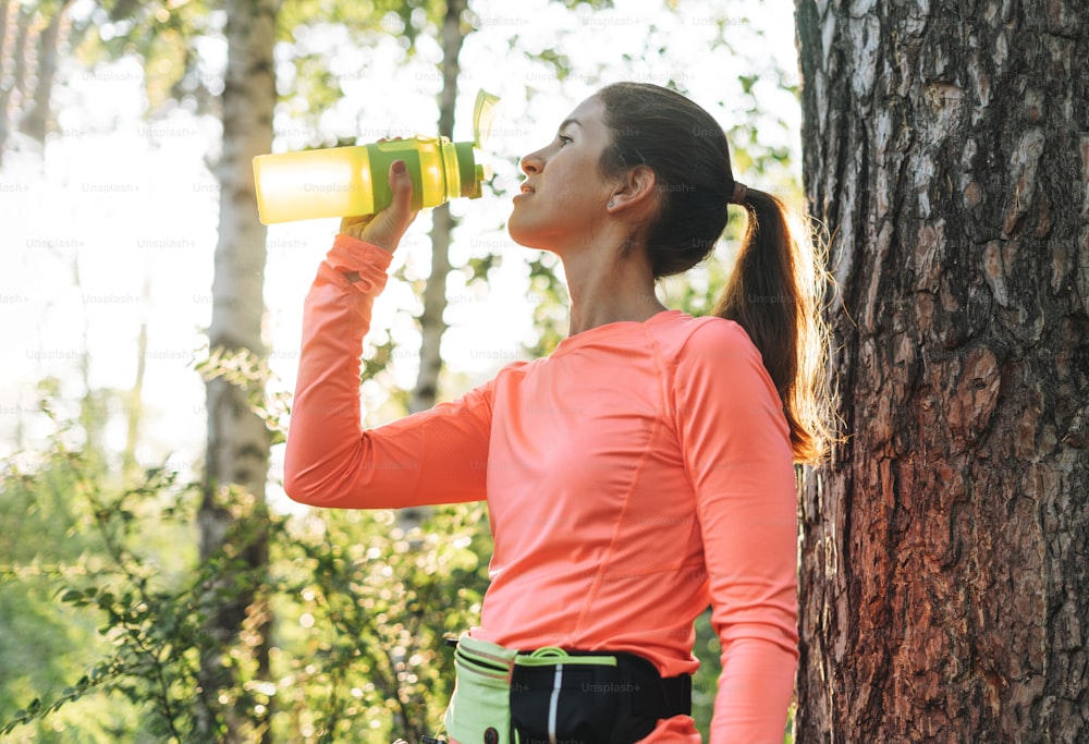 ゴールデンアワーの日の出時間に森でスポーツ服を着て走り、水を飲む若い女性のブルネット。健康とウェルネス、フィットネスライフスタイル