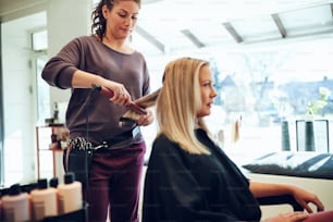머리를 스타일링하는 젊은 미용사 그녀의 살롱에서 일하는 동안 스트레이트너가 있는 의자에 앉아 있는 여성 고객