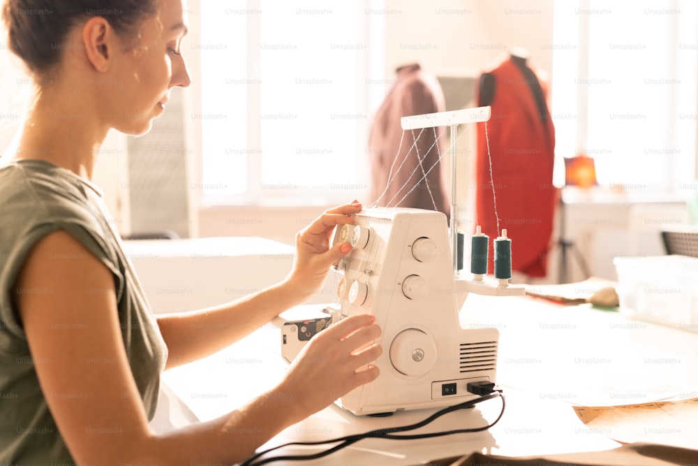 Mujer joven en ropa casual sentada junto a la máquina de coser eléctrica y preparándola para el trabajo