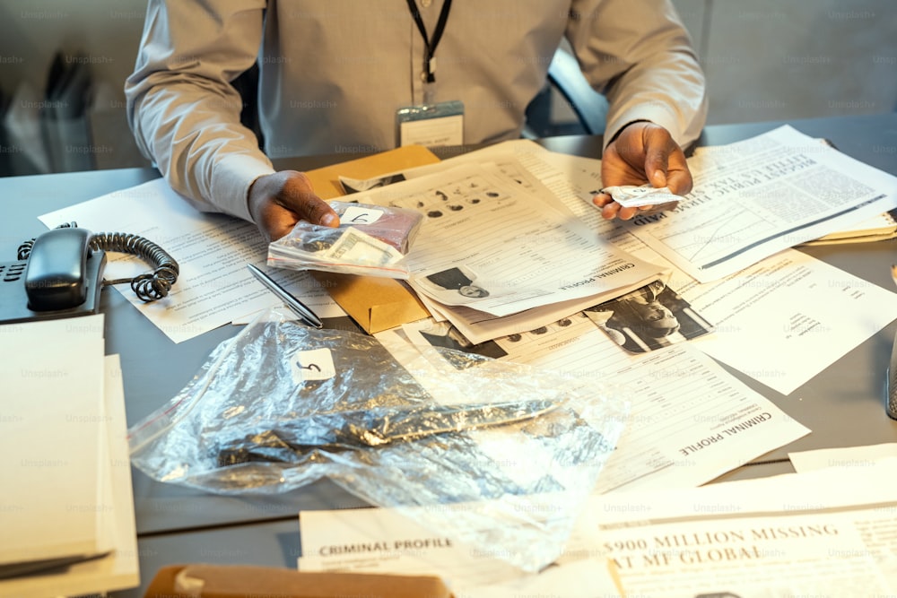 Hands of young contemporary FBI agent or detective holding evidences in packets while sitting by workplace with criminal profiles