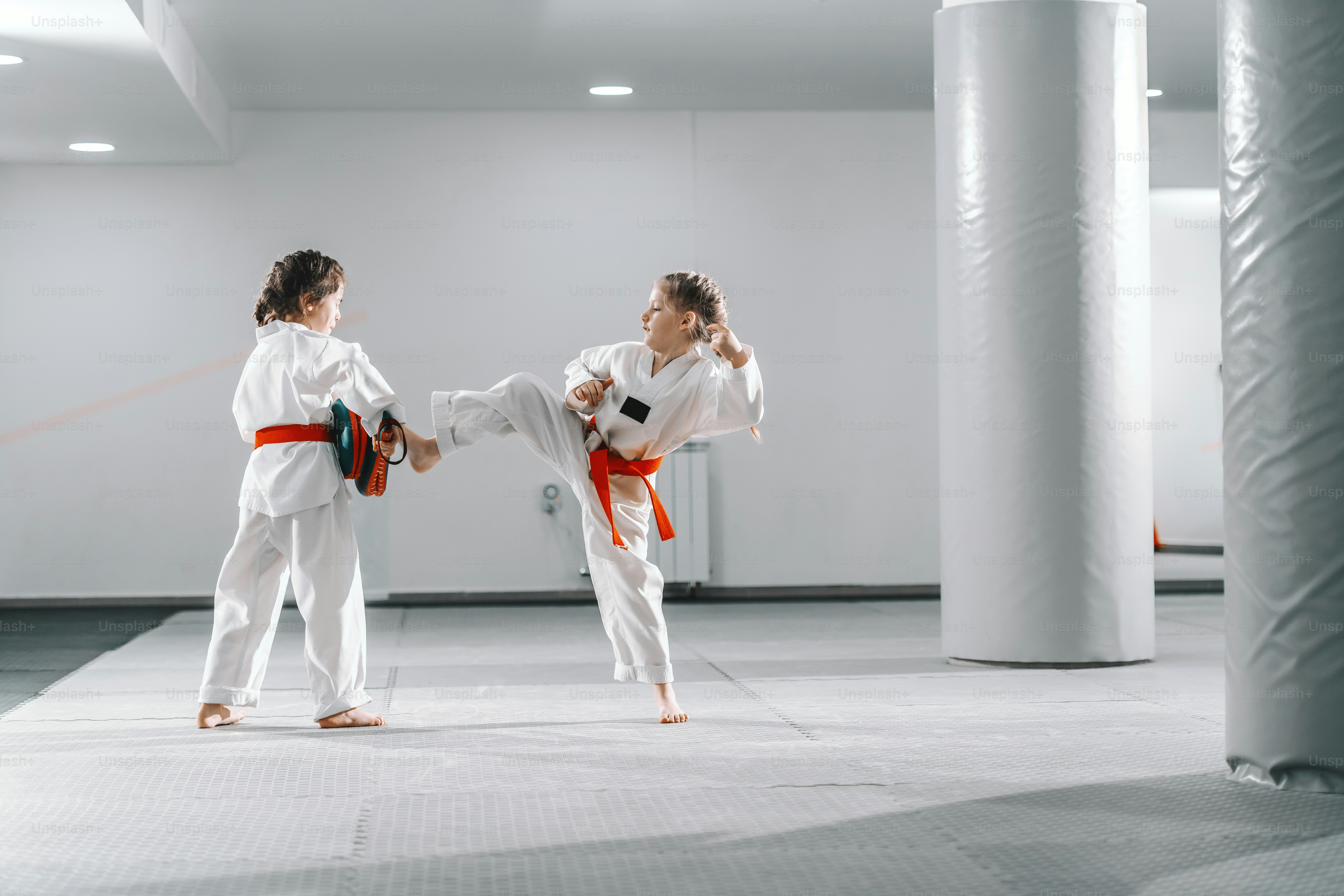 Duas jovens caucasianas em doboks tendo treinamento de taekwondo na academia. Uma menina chutando enquanto outra segurava o chute no alvo.