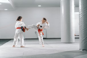 Deux jeunes filles caucasiennes en doboks s’entraînent au taekwondo au gymnase. Une fille donne des coups de pied tandis que l’autre tient une cible de coup de pied.