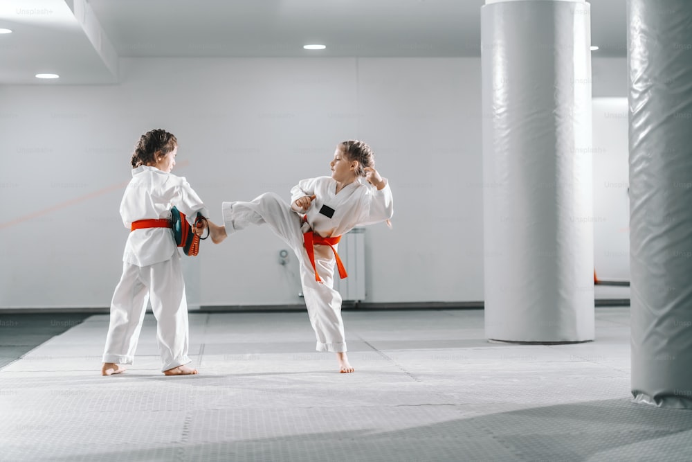 Zwei junge kaukasische Mädchen in Doboks beim Taekwondo-Training im Fitnessstudio. Ein Mädchen tritt, während ein anderes das Ziel hält.